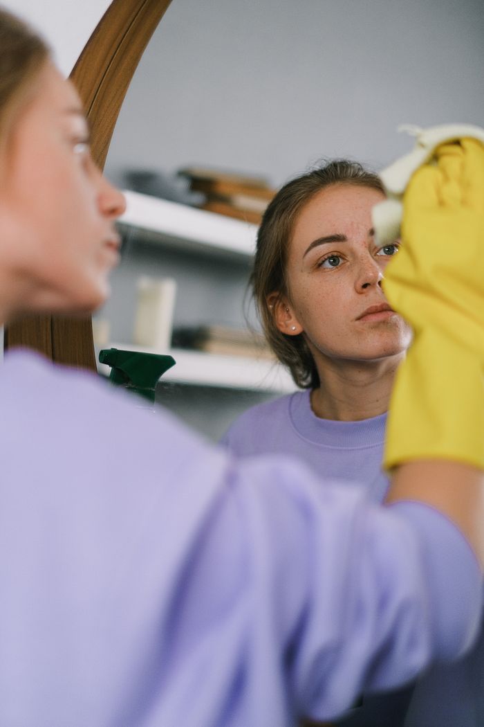 Female crop shot in a purple sweatshirt and latex gloves cleaning a mirror in a bright apartment during the day